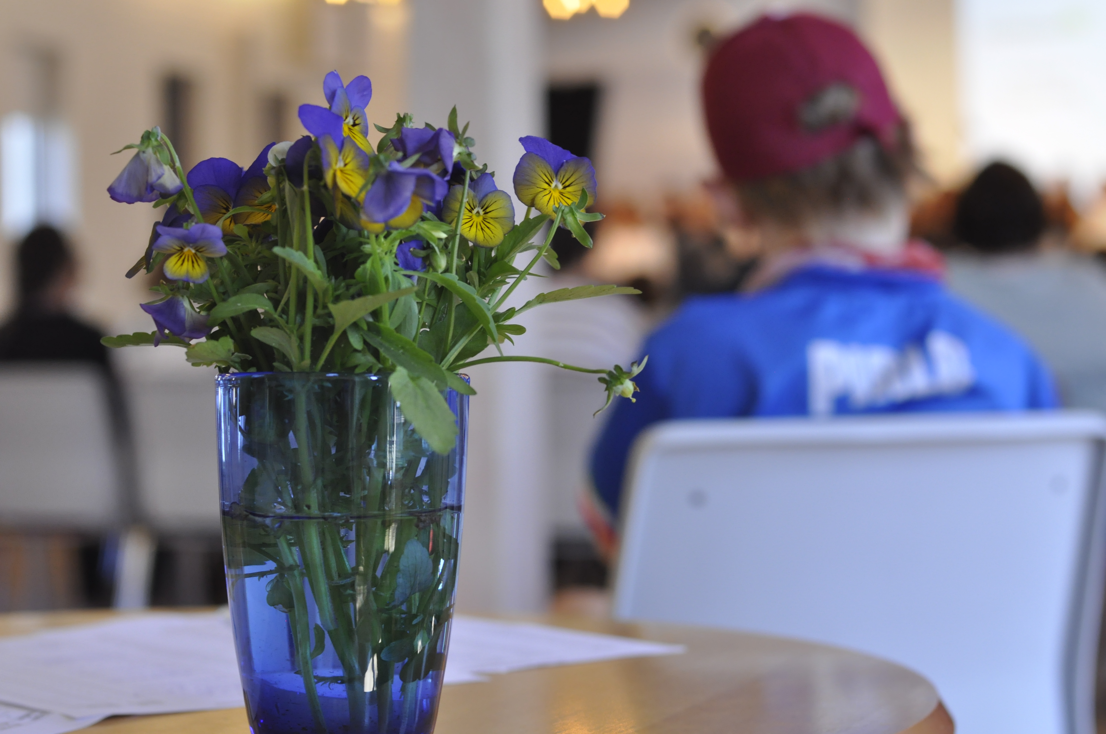 En bukett med lila och gula blommor i en vas och i bakgrunden en personer med blå tröja