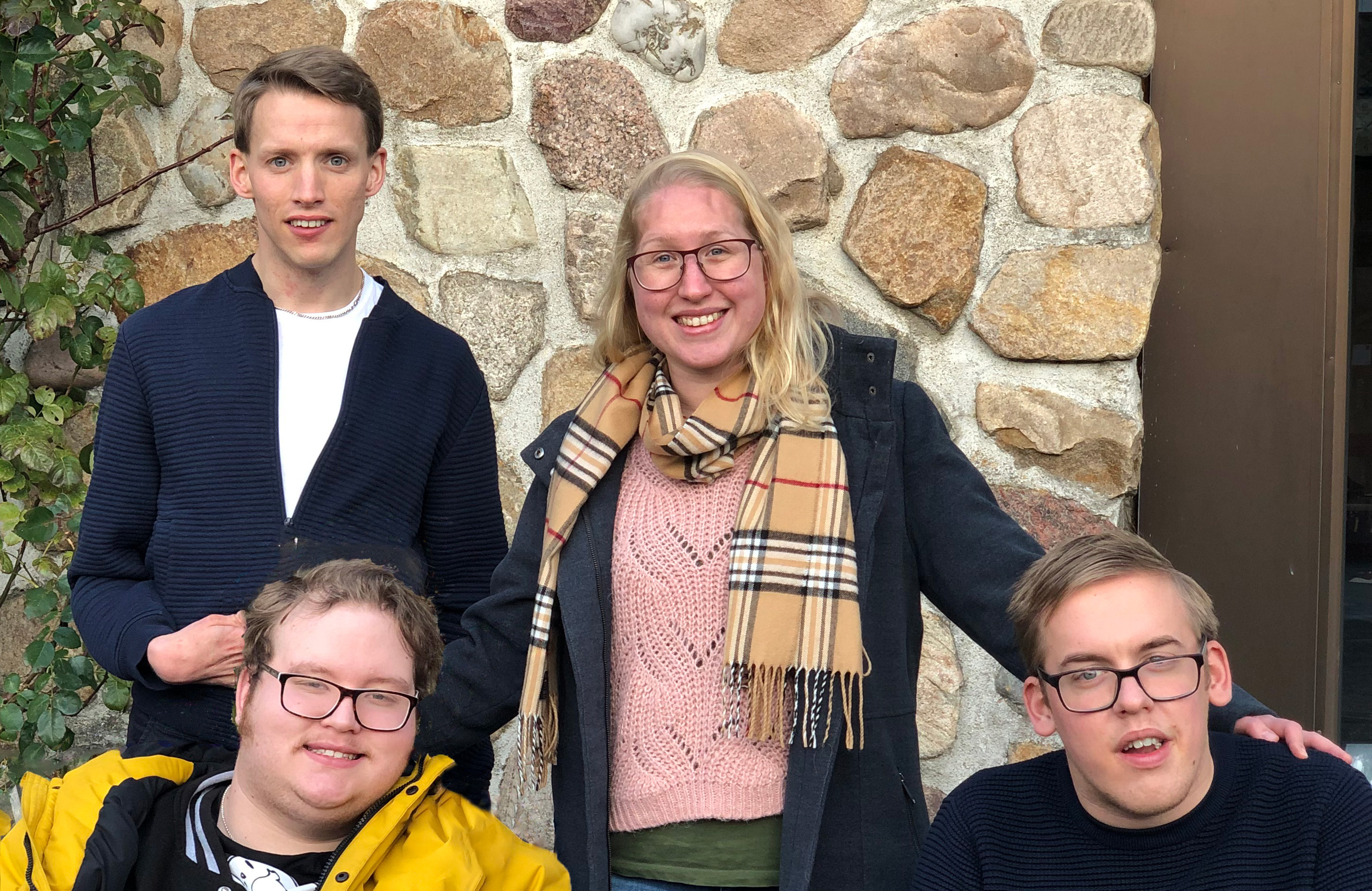 Två personer som sitter och två som står framför en stenvägg utomhus.